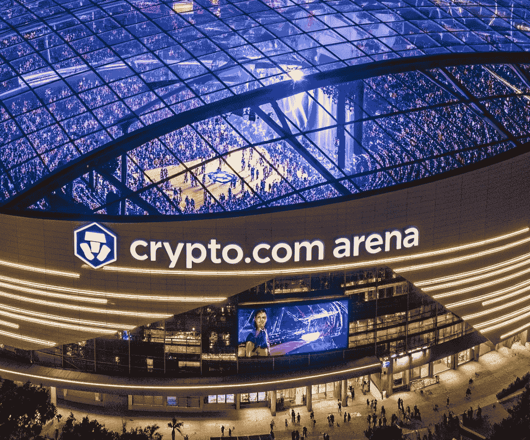 An aerial, nighttime view of the Crypto.com Arena in downtown Los Angeles, with a large crowd visible inside. The city skyline is brightly lit in the background.